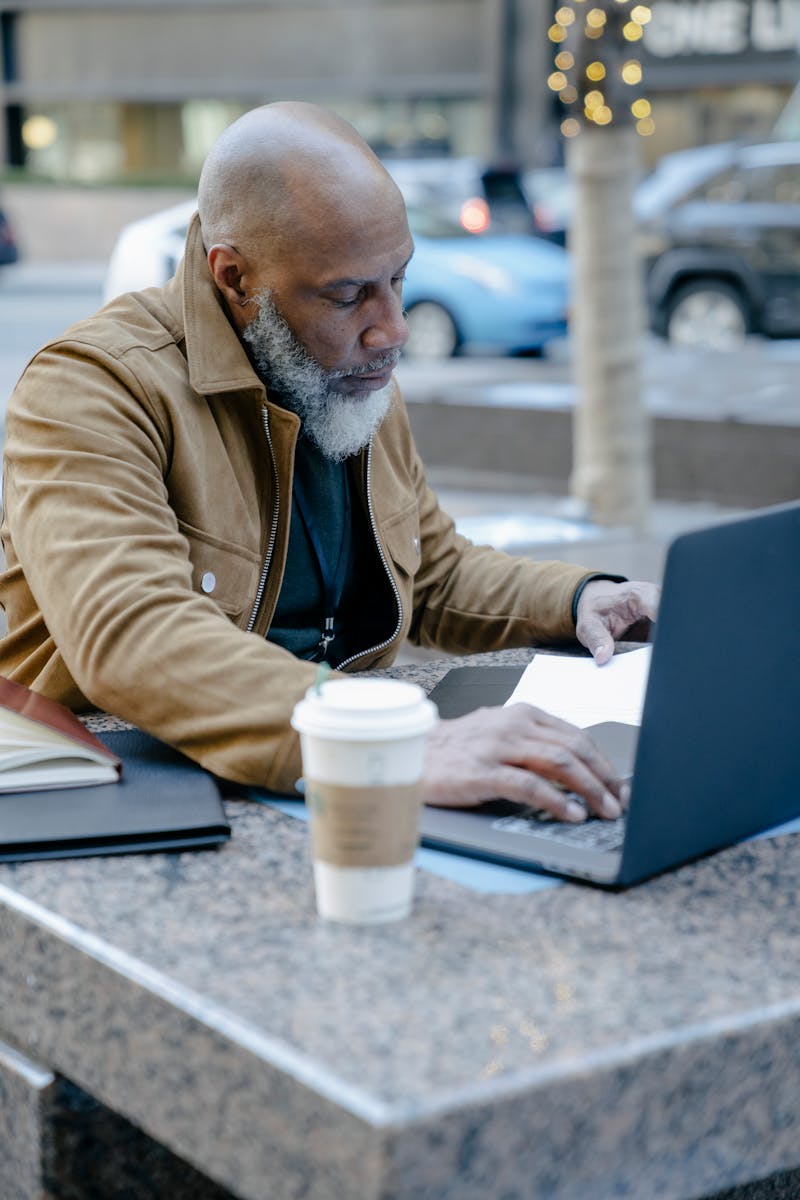 Bald man in a brown jacket using a laptop outdoors with a coffee cup on a marble table.